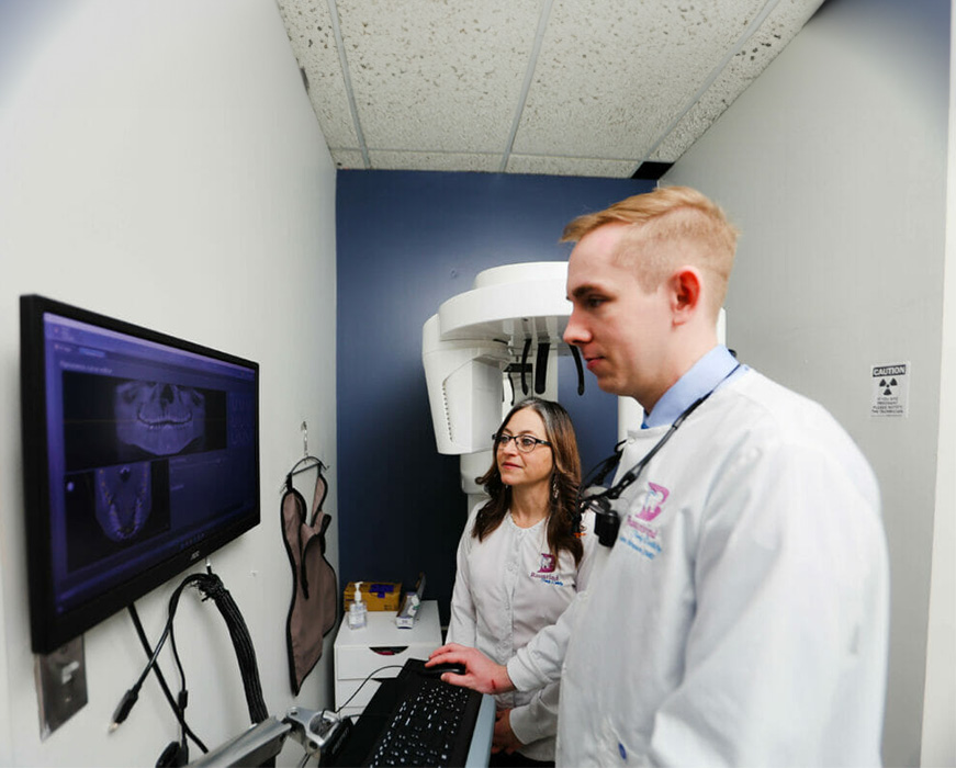 A dentist and dental hygienist are standing in front of a dental X-ray machine, examining an image on the screen.