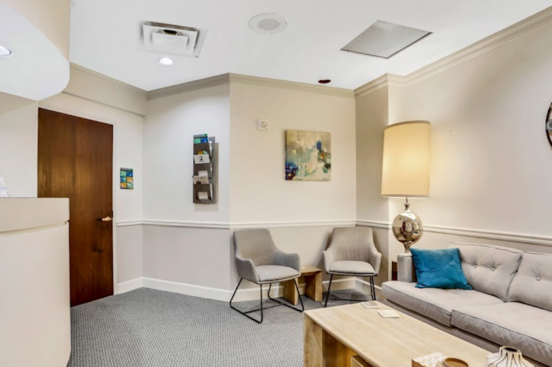 The image shows an interior space that appears to be a waiting area or reception room, featuring a reception desk with a chair and a lamp, a wall-mounted art piece, and a modern lobby with a seating arrangement.