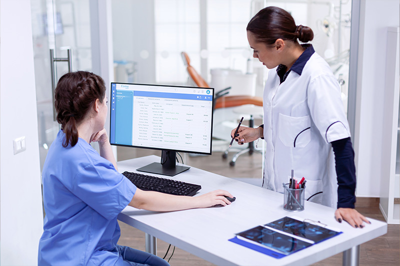 A healthcare professional, possibly a nurse or doctor, is standing behind a desk with a laptop and monitor, while another individual is seated at the desk. They are both in an office setting, which appears to be a modern medical facility.