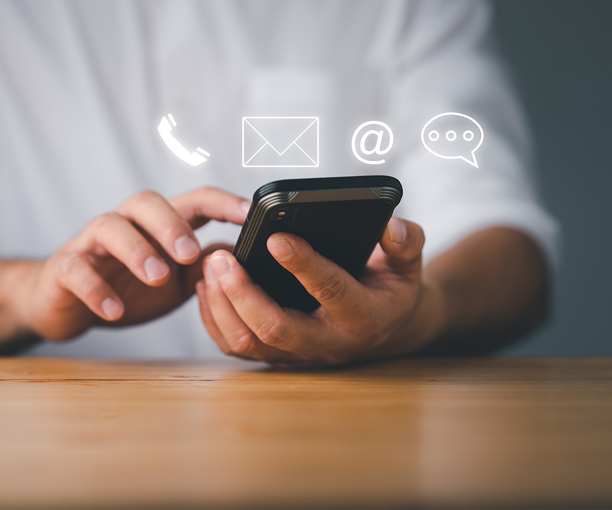 The image shows a person s hand holding a smartphone with various icons displayed on the screen, including an envelope, a phone, and a speech bubble. The individual appears to be using the device, possibly texting or browsing. The background is blurred but suggests an indoor setting.